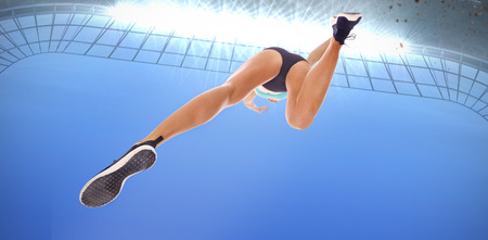 Low angle female athlete jumping against large football stadium under bright blue skyの写真素材
