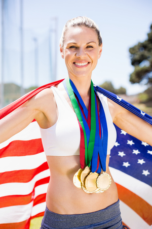Portrait of happy female athlete holding up american flag with gold medal in stadiumの写真素材
