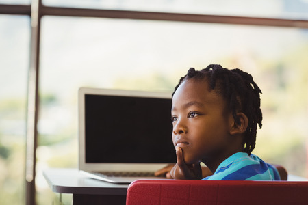 Thoughtful schoolboy sitting on chair and using laptop at school