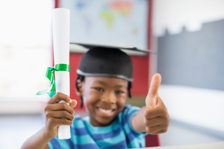 Portrait of schoolboy in mortar board holding certificate and showing thumbs up in classroom at schoolの写真素材