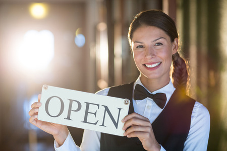 Portrait of smiling waitress holding open sign in restaurantの写真素材