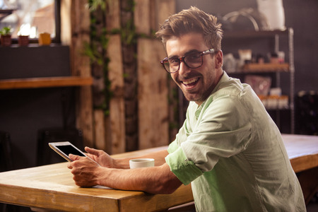 Happy hipster man posing for camera while using tablet at cafeの写真素材