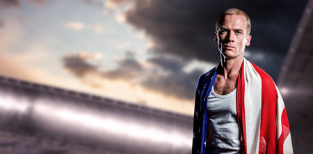 Athlete with american flag wrapped around his body against composite image of arena and cloudy skyの写真素材