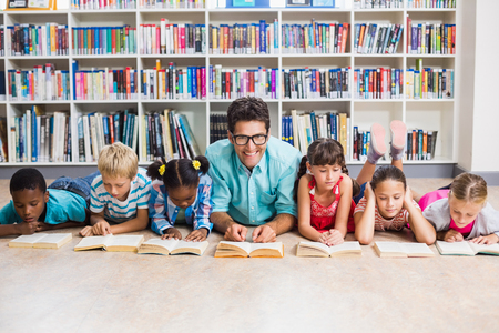 Teacher and kids lying on floor reading book in libraryの写真素材