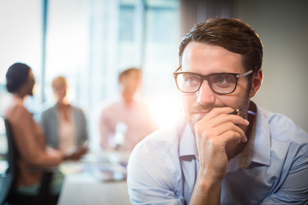 Thoughtful man with hand on chin while coworker interacting in the background in the officeの写真素材