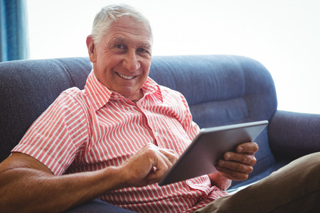 Senior man seated on a sofa looking at camera while holding digital tabletの写真素材