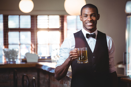 Portrait of smiling bartender standing at bar counter offering a glass of beerの写真素材