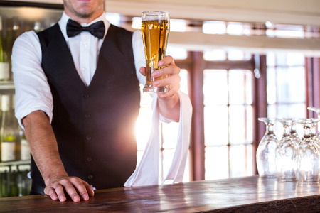 Mid section of bartender standing at bar counter offering a glass of beer