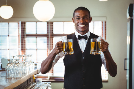 Portrait of smiling bartender standing at bar counter offering a two glass of beer