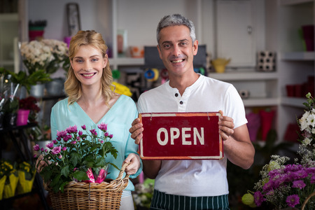 Portrait of smiling florists holding open sign placard and flower basket in flower shop