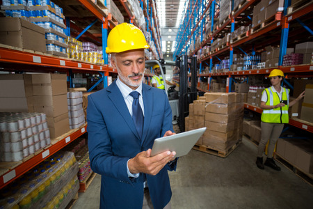 Focus of manager is using a tablet and wearing a hard hat in a warehouse