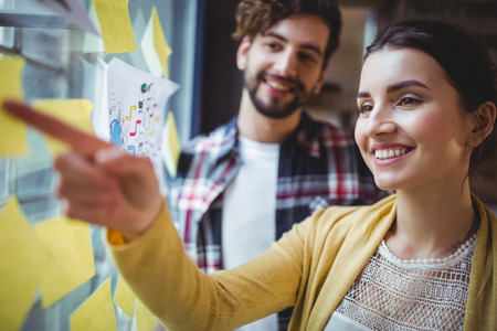 Happy businesswoman pointing at sticky notes while explaining male colleague in creative officeの写真素材