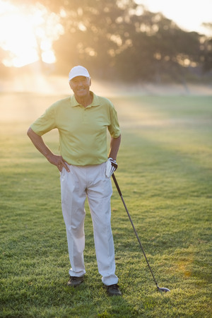 Portrait of smiling golfer with hand on hip while holding golf club on fieldの写真素材