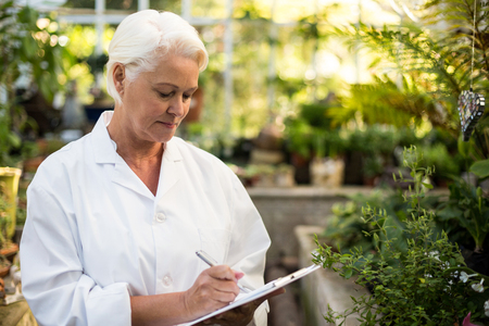 Mature female scientist writing in clipboard while standing at greenhouseの写真素材