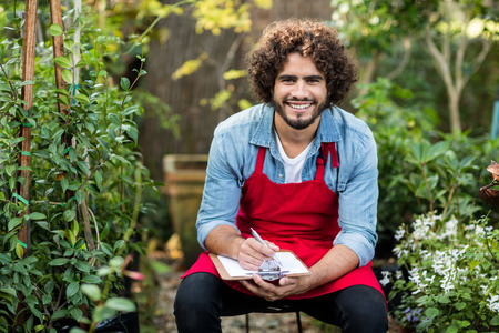 Portrait of happy male gardener writing on clipboard while sitting at greenhouse