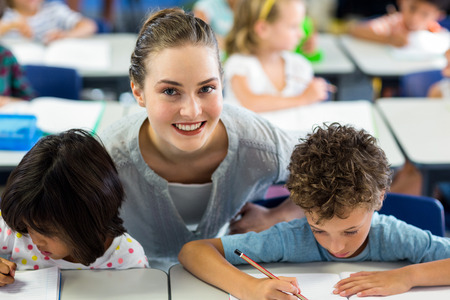 Portrait of happy female teacher helping schoolchildren to write on bookの写真素材