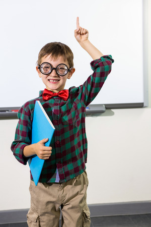 Portrait of cute boy with notebook raising hand against whiteboard in classroomの写真素材
