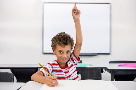 Portrait of elementary boy raising hand against whiteboard in classroom