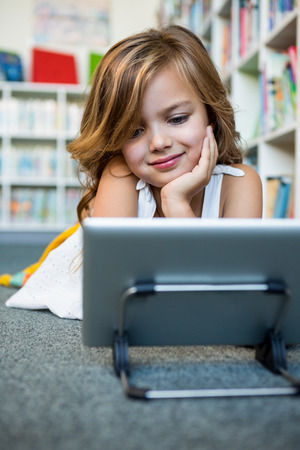Smiling girl using digital tablet while lying in school libraryの写真素材