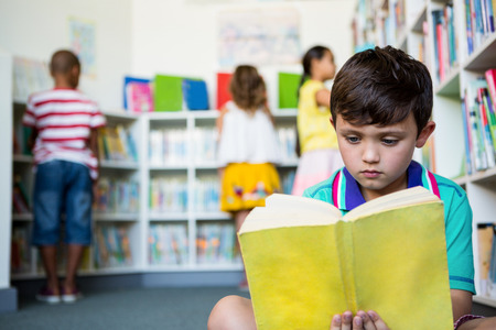 Elementary boy reading book with students in background at school libraryの写真素材