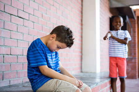 Schoolboy laughing on sad classmate in corridor at schoolの写真素材