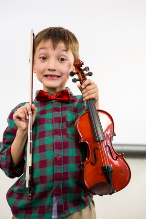 Portrait of cute boy holding violin with bow in classroomの写真素材