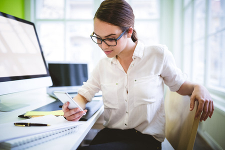 Female graphic designer using phone at desk in creative officeの写真素材