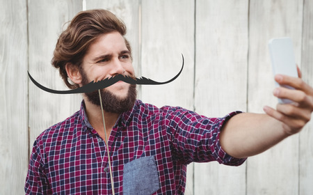 Hipster taking selfie against wooden wall against artificial mustache