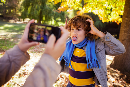 Cropped image of girl photographing boy making face in park during autumnの写真素材