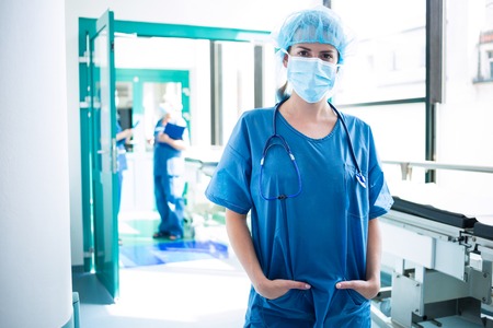 Portrait of female surgeon standing with hands in pockets in corridor at hospital