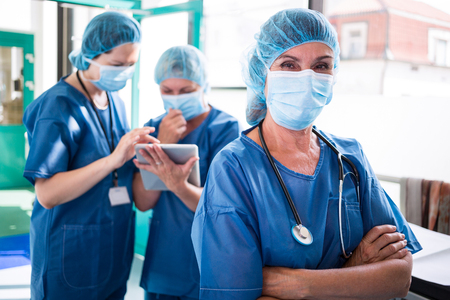 Portrait of surgeon standing with arms crossed while staff discussing over digital tablet in background at hospitalの写真素材
