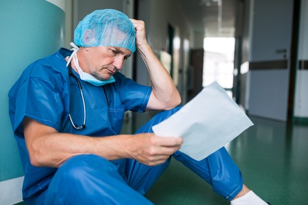 Sad surgeon sitting on floor with report in corridor at hospitalの写真素材