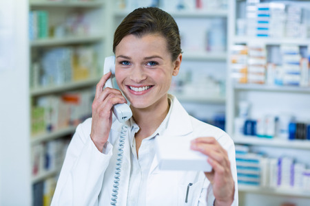 Portrait of pharmacist holding medicine box while talking on phone in pharmacyの写真素材