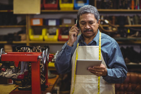 Shoemaker using digital tablet while talking on mobile phone in workshopの写真素材
