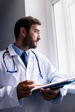 Thoughtful doctor looking through window while holding clipboard in hospitalの写真素材
