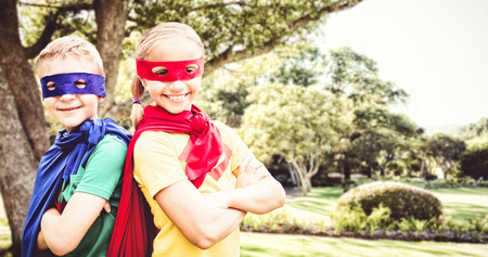 Happy brother and sister in cape and eye mask against bench by tree in park