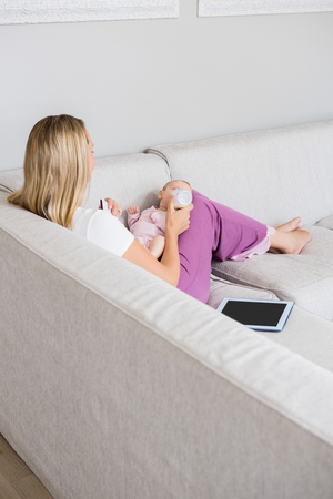 Mother feeding baby with milk bottle in living room at homeの写真素材