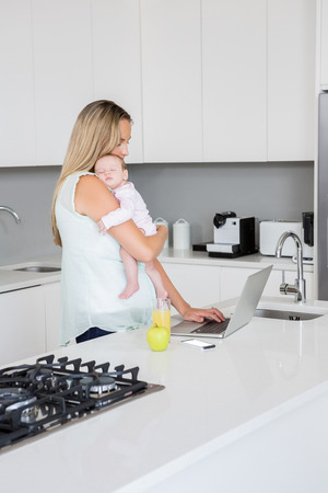 Mother using laptop while carrying baby in kitchen at homeの写真素材