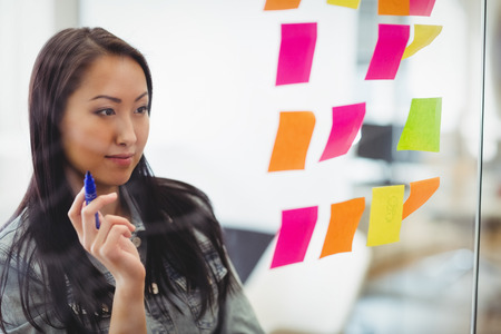 Confident creative businesswoman looking at multi colored sticky notes on glass in meeting room at...