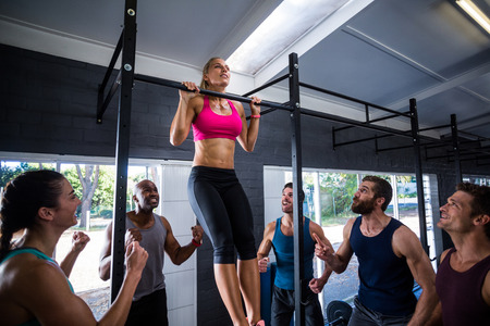 Happy friends cheering woman doing chin-ups in gym