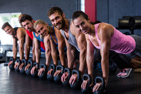 Portrait of smiling friends doing push-ups with kettlebell while exercising in gym