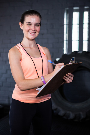 Portrait of smiling female fitness instructor writing in clipboard while standing in gymの写真素材