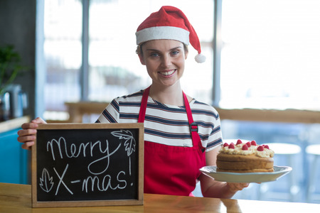 Portrait of smiling waitress holding slate with merry x-mas sign and cake in cafÃ©の写真素材