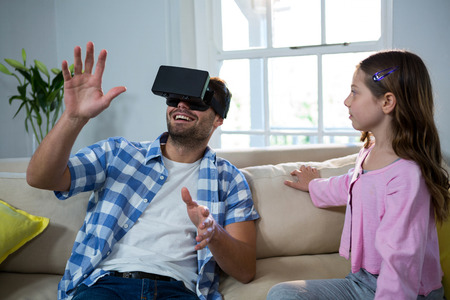 Father using virtual reality headset while daughter sitting beside him in the living room at homeの写真素材