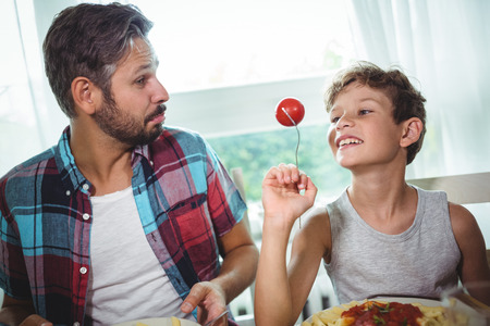 Smiling boy showing a cherry tomato to his father while having mealの写真素材