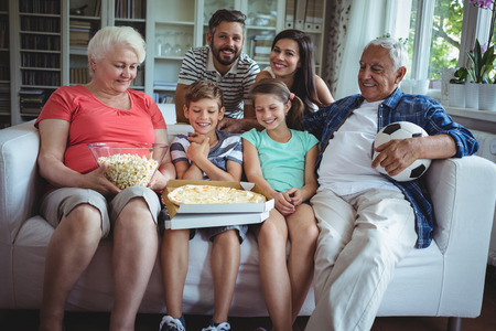 Multi-generation family sitting on sofa with popcorn and pizza while watching soccer match