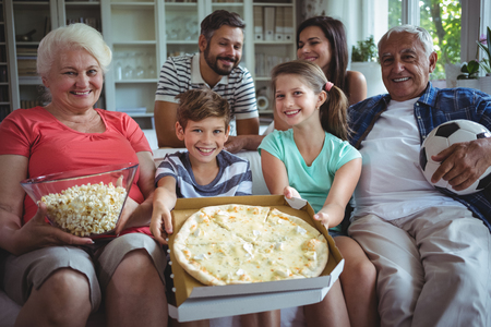 Multi-generation family sitting on sofa with popcorn and pizza while watching soccer match