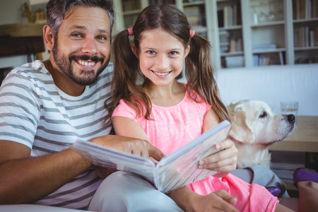 Smiling father and daughter looking at photo album in living room at homeの写真素材