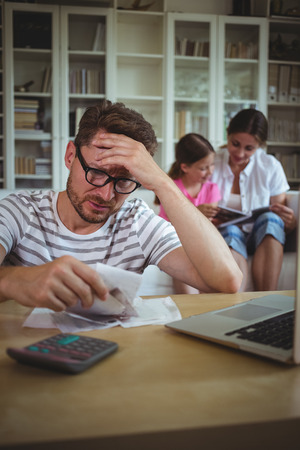 Worried man calculating bills while his wife and daughter sitting on sofa in living roomの写真素材