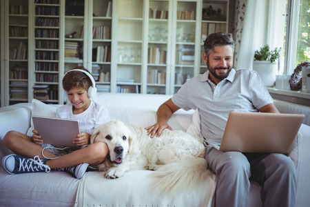 Father and son sitting on sofa and using digital tablet and laptop in living room at homeの写真素材